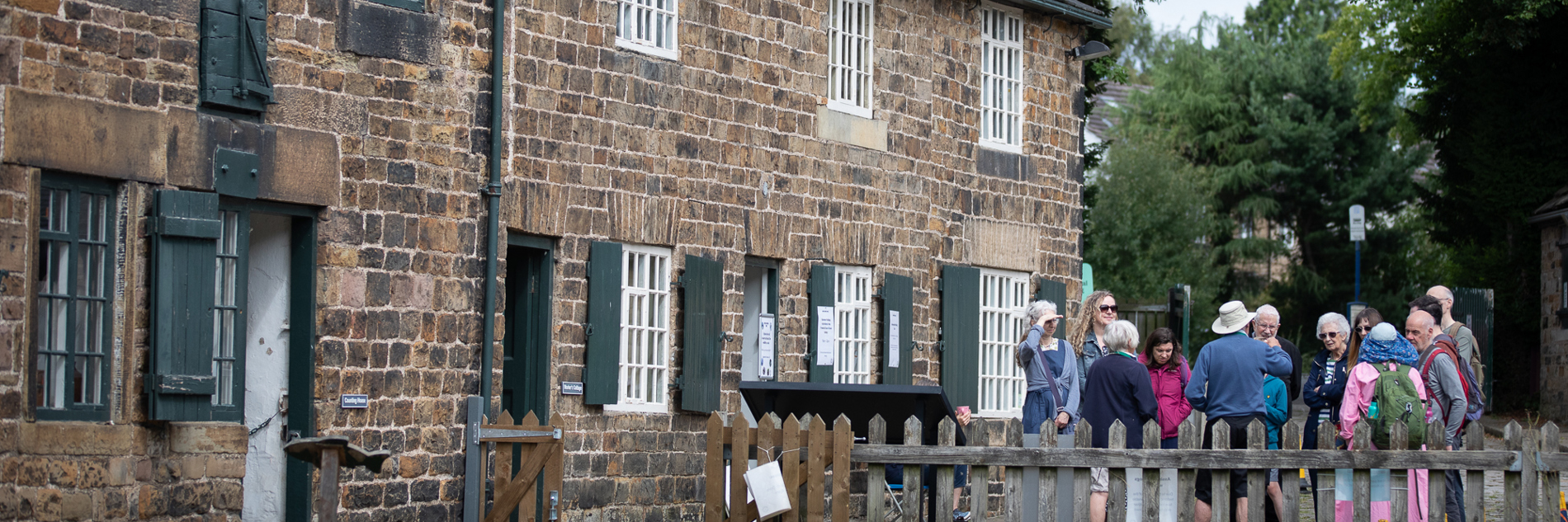 A group visitors gathered in front of a row of historic terraced stone cottage with trees in the background 