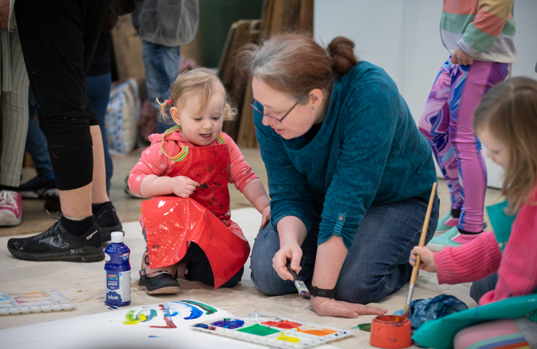 A photograph of a young girl and a woman kneeling on the floor making an image with paintbrushes. They are in conversation and there is a palette in front of them with brightly coloured paints.