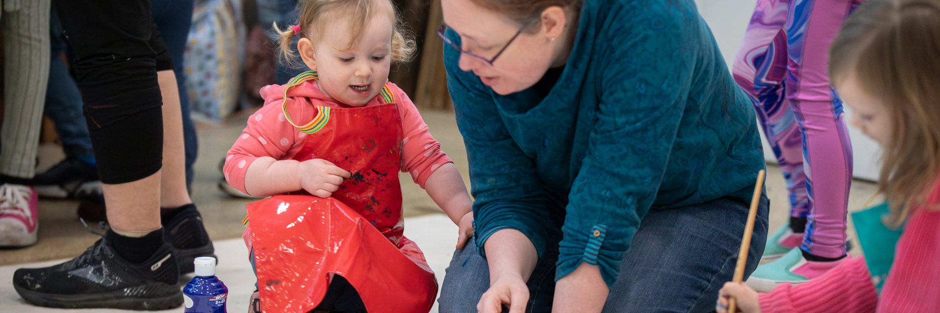 A photograph of a young girl and a woman kneeling on the floor making an image with paintbrushes. They are in conversation and there is a palette in front of them with brightly coloured paints.