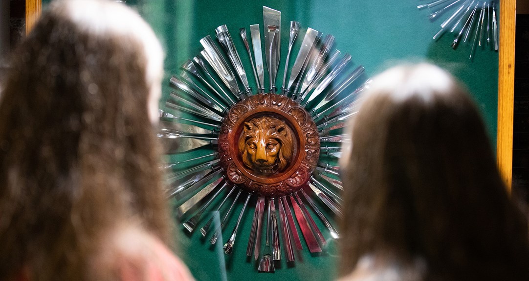 The back of two people's head as they look at a round wooden lion carving with metal tools arranged around it like sun rays.