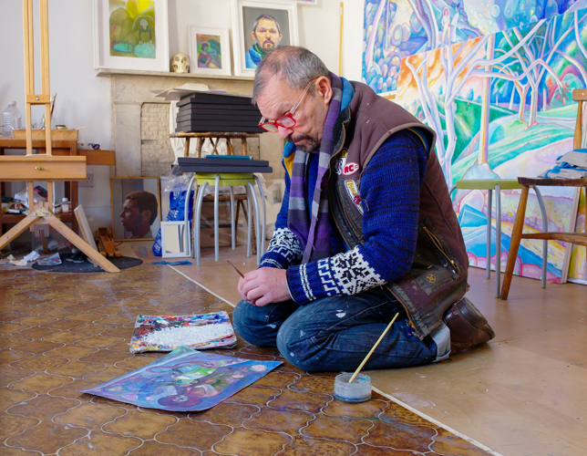 A photograph of an adult sitting on a wooden and lino floor and looking at an artwork. They have a paintbrush in their hand and a pot and paintbrush next to them. In the background there is an easel, a table with art supplies, and colourful artworks propped up again the wall and fireplace. 