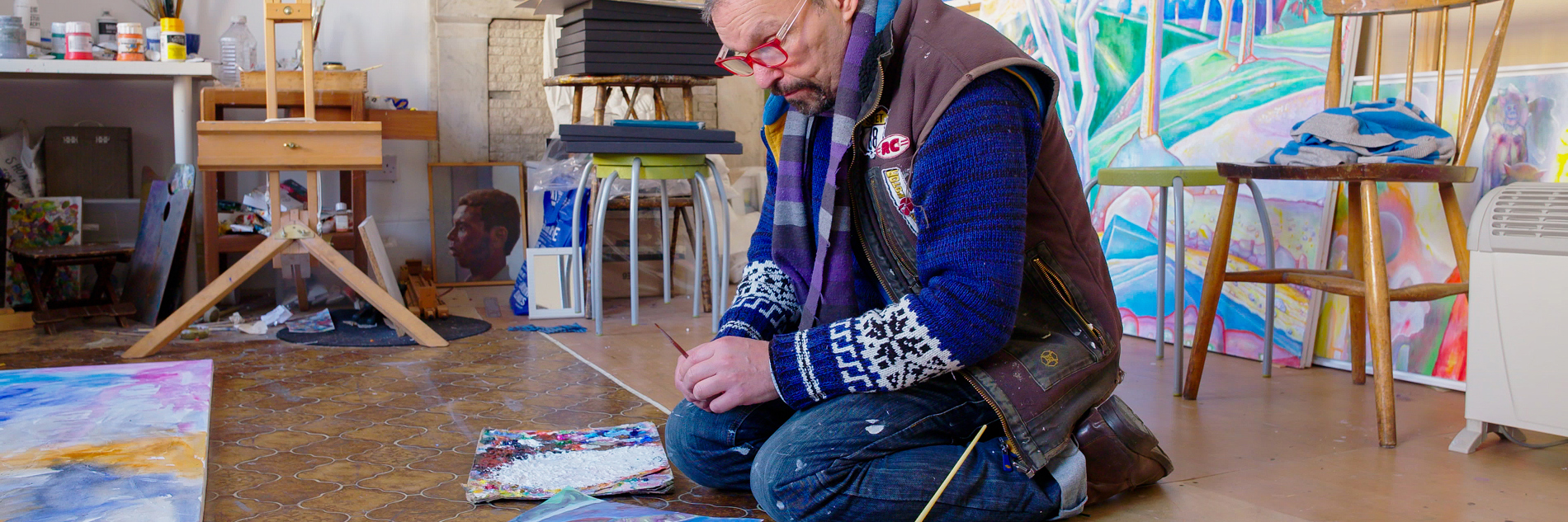 A photograph of an adult sitting on a wooden and lino floor and looking at an artwork. They have a paintbrush in their hand and a pot and paintbrush next to them. In the background there is an easel, a table with art supplies, and colourful artworks propped up again the wall and fireplace. 