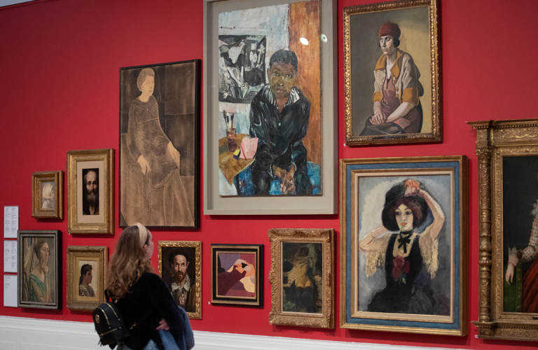 A visitor with long hair and a black top looks at display of a group of portrait artworks. The artworks are hung closely together on a red wall.
