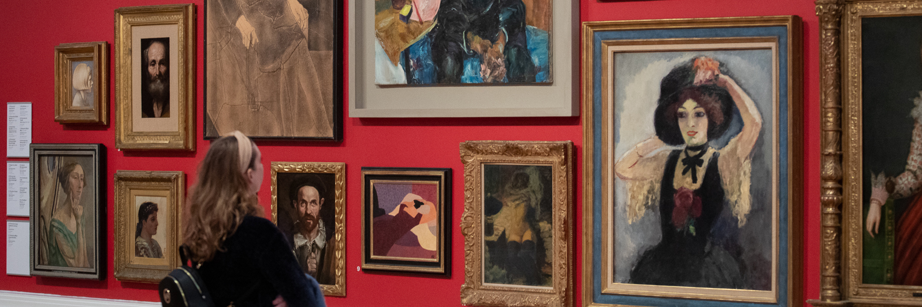 A visitor with long hair and a black top looks at display of a group of portrait artworks. The artworks are hung closely together on a red wall.