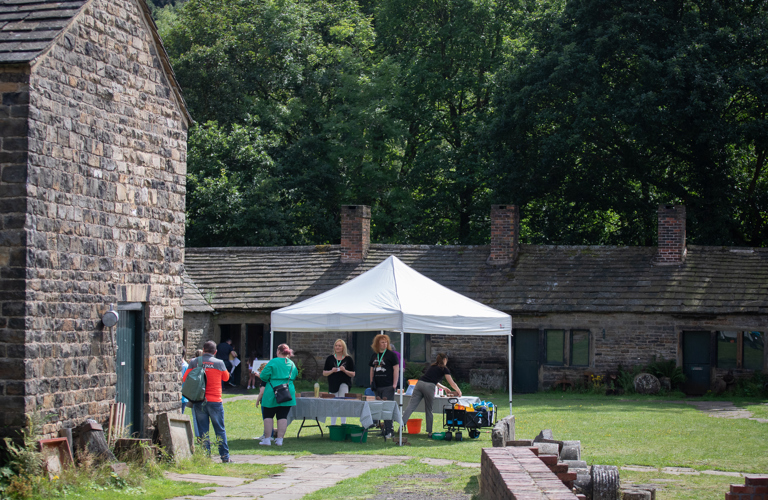 Staff and visitors around a marquee, which is placed on a green lawn in front of a row of single storey historic stone buildings