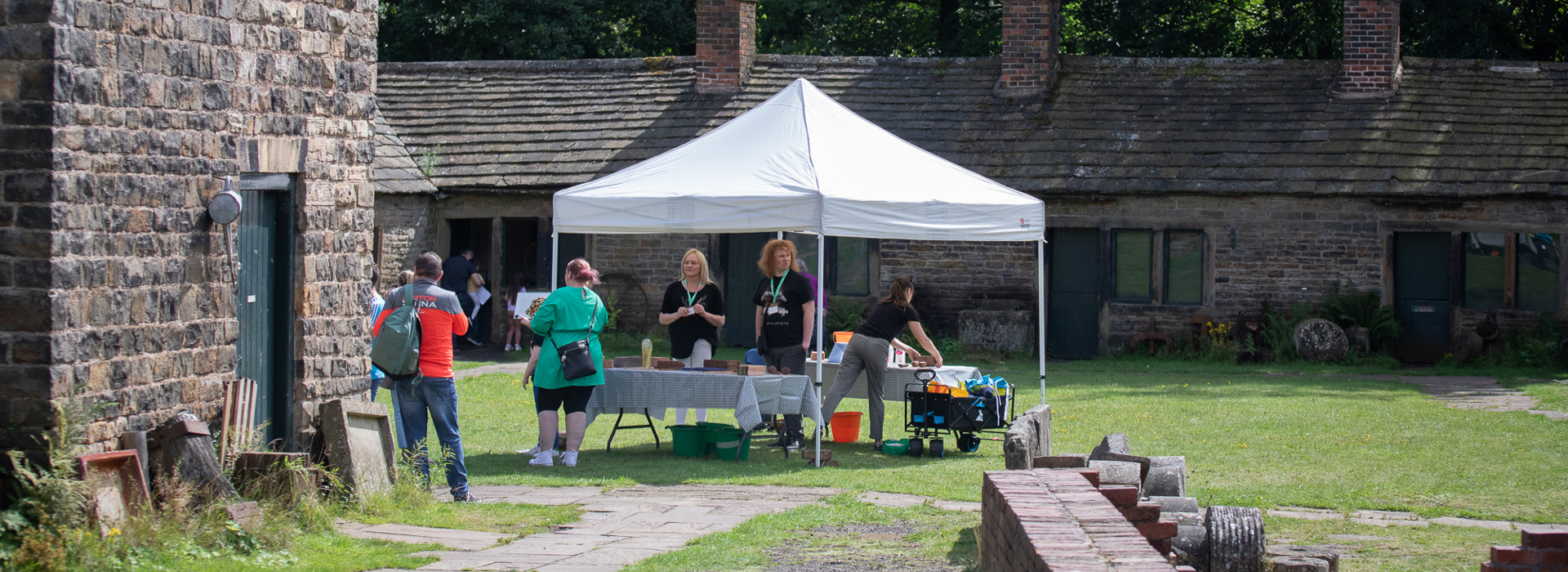 Staff and visitors around a marquee, which is placed on a green lawn in front of a row of single storey historic stone buildings