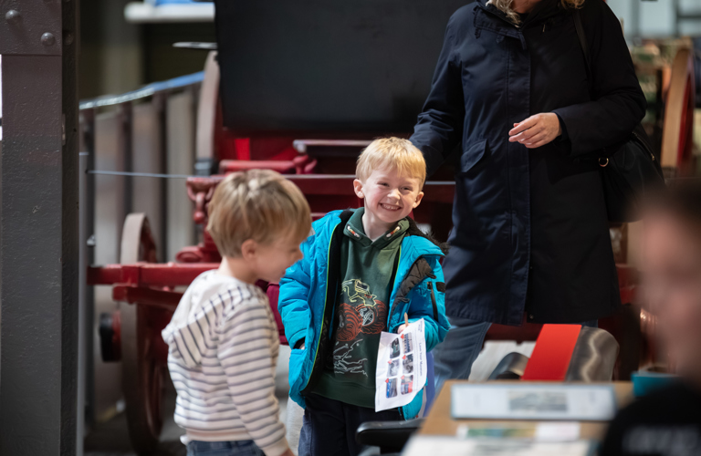 A photograph of two young boys smiling at Kelham Island Museum. The red frame of a carriage is visible behind them