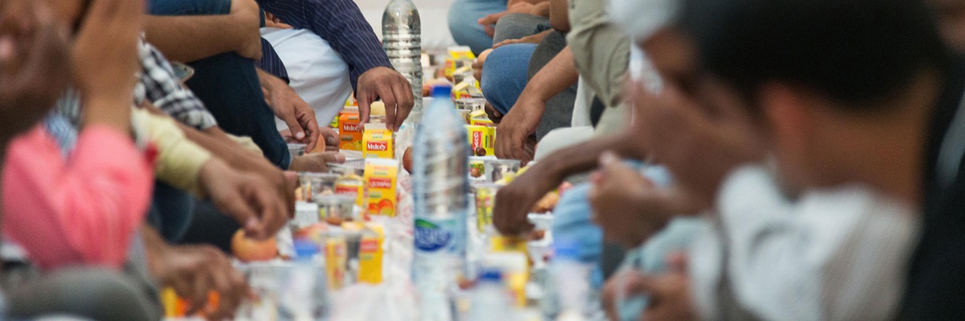 A photograph showing people gathered around a table sharing food and drink. There are large plastic bottles of water, little yellow cartons of drink, and plates of food. Everyone is at cross-legged on the floor.