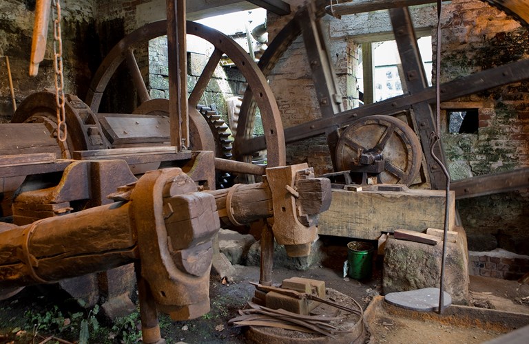 Two rusting tilt forge hammers mounted on huge old timber shafts with gearwheel drive mechanisms in the background.