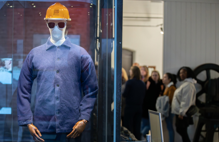 A photograph of a mannequin wearing a blue boiler suit and a yellow hardhat