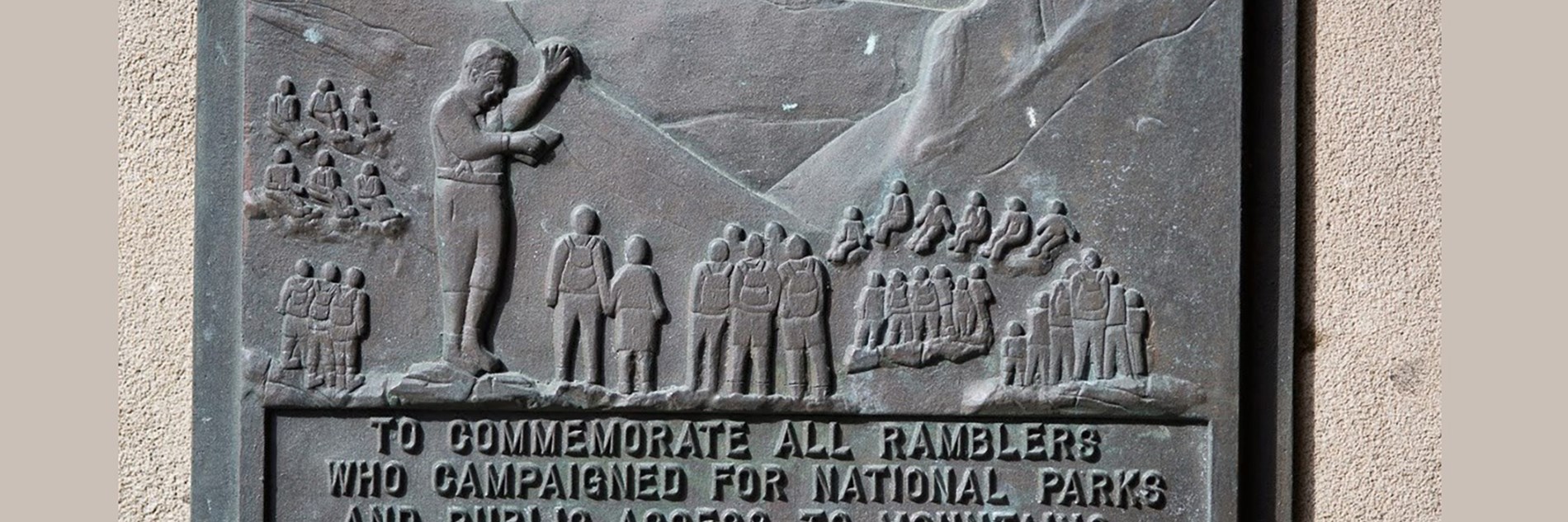 A commemorative stone relief plaque showing groups of ramblers walking and sitting, enjoying the countryside. There are hills in the background. The inscription reads "To commemorate all ramblers who campaigned for National Parks and public access to mountains and moorland. Year 2000".
