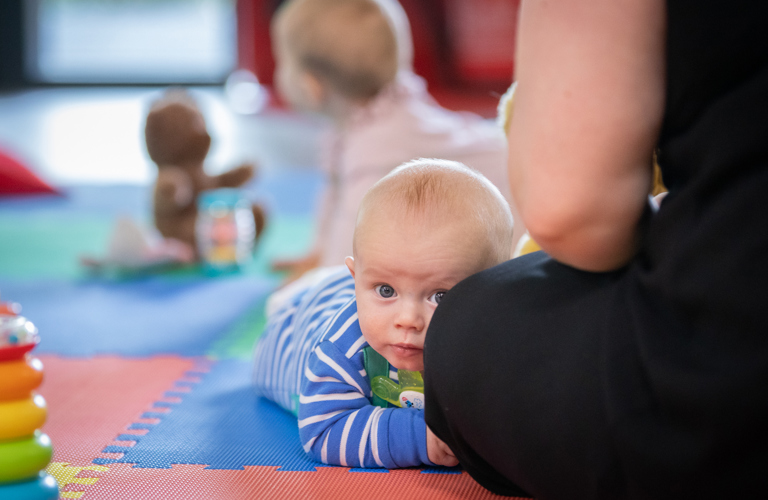A photograph of a baby lying on their front looking at the camera. An adult is sitting next to the baby and in the background there is another baby and some toys. 
