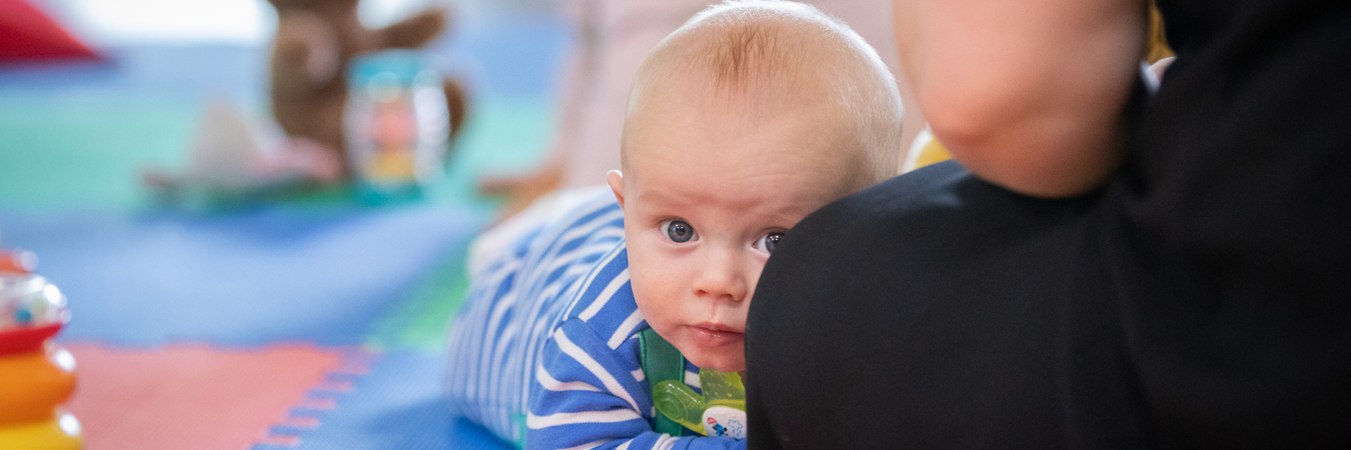A photograph of a baby lying on their front looking at the camera. An adult is sitting next to the baby and in the background there is another baby and some toys. 
