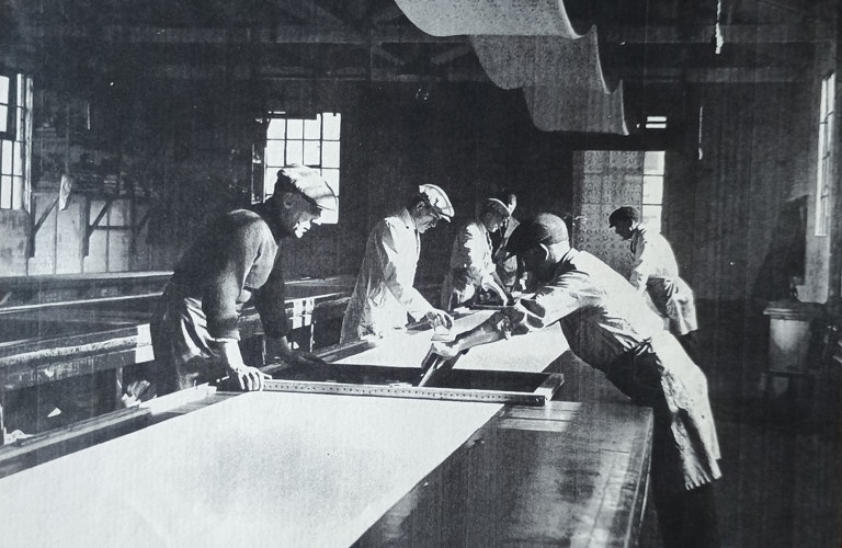 A black and white photo of workers in a screen printing factory around 1935. Six men are working around a long wooden table with metres of material stretched on top of it.