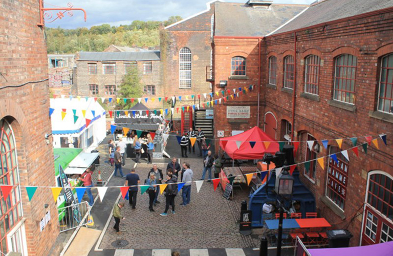 Photograph of a courtyard, taken from above on a sunny day. There are red brick buildings, multicoloured bunting and people walking amongst stalls