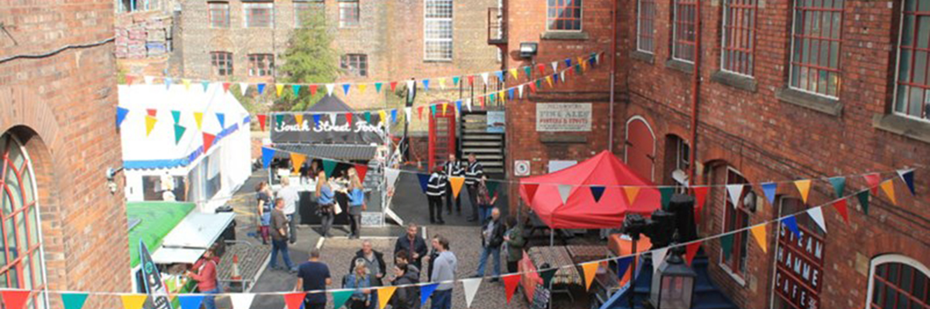 Photograph of a courtyard, taken from above on a sunny day. There are red brick buildings, multicoloured bunting and people walking amongst stalls