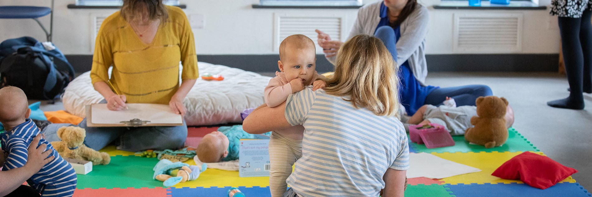 A group of babies and parents, sat on colourful mats, surrounded by baby toys and cushions. Some of the adults are drawing on large clipboards.
