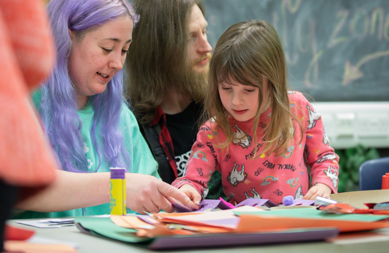 A photograph of a small child being assisted in a crafting activity by an adult with purple hair. There is a lots of paper and a gluestick on the table in front of them.