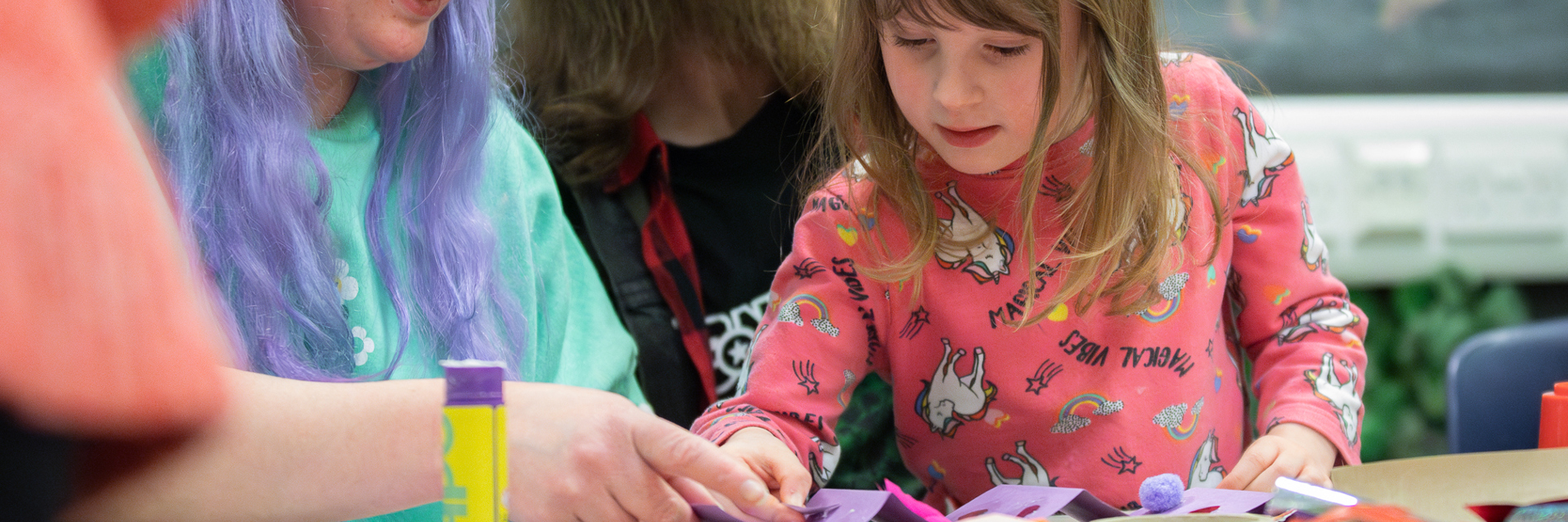 A photograph of a small child being assisted in a crafting activity by an adult with purple hair. There is a lots of paper and a gluestick on the table in front of them.