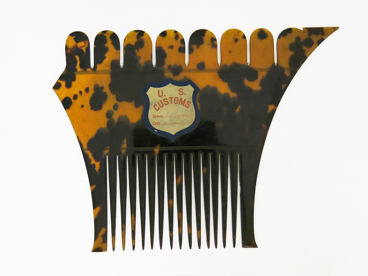 An unusually shaped comb with scalloped edging along the top and curved sides. It has a tortoise shell pattern and a faded sticker on the front that says 'U.S Customs' on it.