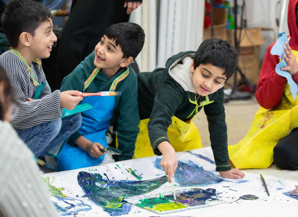 Children smile and chat as they kneel on teh floor and contribute to a large-scale artwork