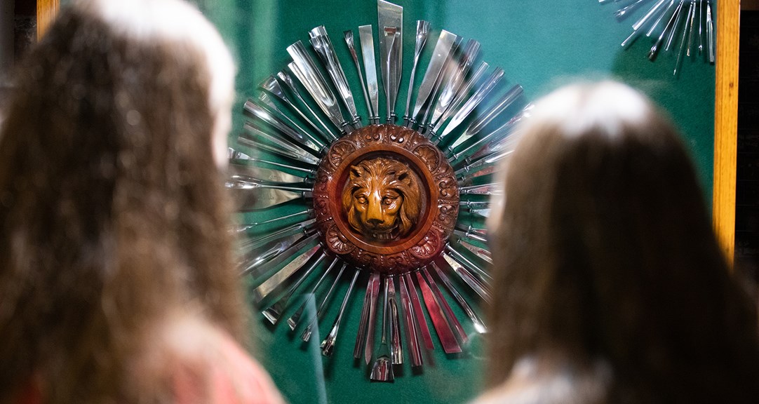 The back of two people's head as they look at a wooden lion carving surrounded by metal tools arranged around it like sun rays.