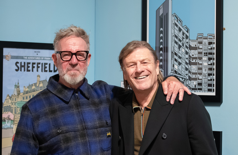 Two men stand arm in arm in front of paintings of Sheffield in an art gallery