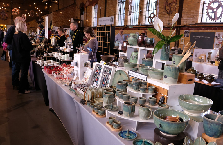 Photograph of a stall inside a large brick hall with windows. The stall features a range of ceramic bowls and cups in shades of blue and green. In the distance, a group of people are talking across the stalls.