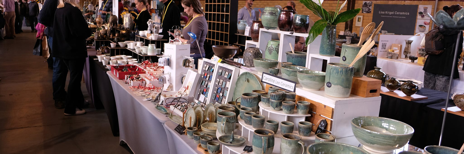 Photograph of a stall inside a large brick hall with windows. The stall features a range of ceramic bowls and cups in shades of blue and green. In the distance, a group of people are talking across the stalls.