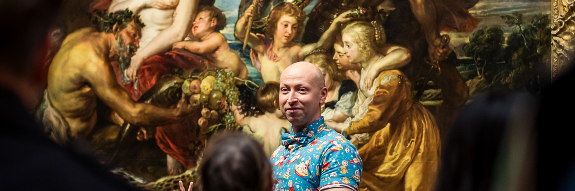 A photograph of a man stood in front of a large painting that seems to depict feasting and war. He is wearing a bright blue patterned shirt and is giving a talk to a crowd of people.