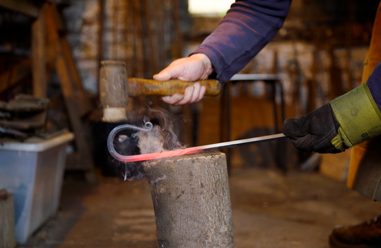 A photograph of arms and hands of a person holding a hammer and a hooked piece of metal in a workshop. The person is wearing a glove on one hand and in the background there are tools and materials.  