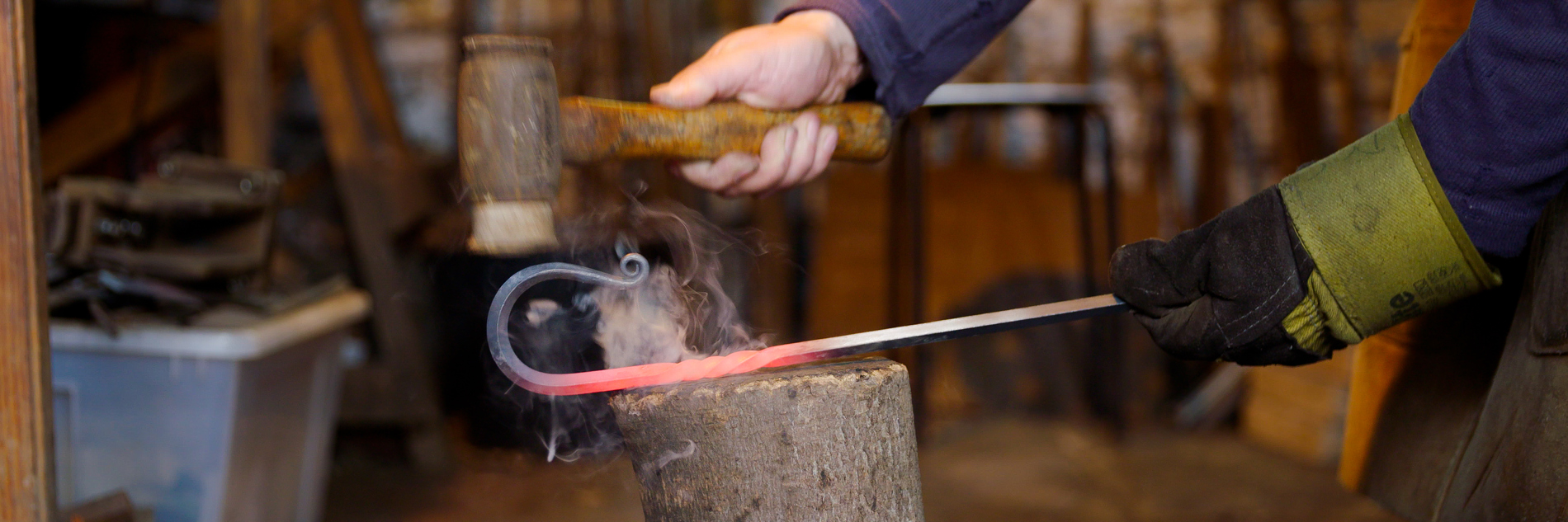 A photograph of arms and hands of a person holding a hammer and a hooked piece of metal in a workshop. The person is wearing a glove on one hand and in the background there are tools and materials.  