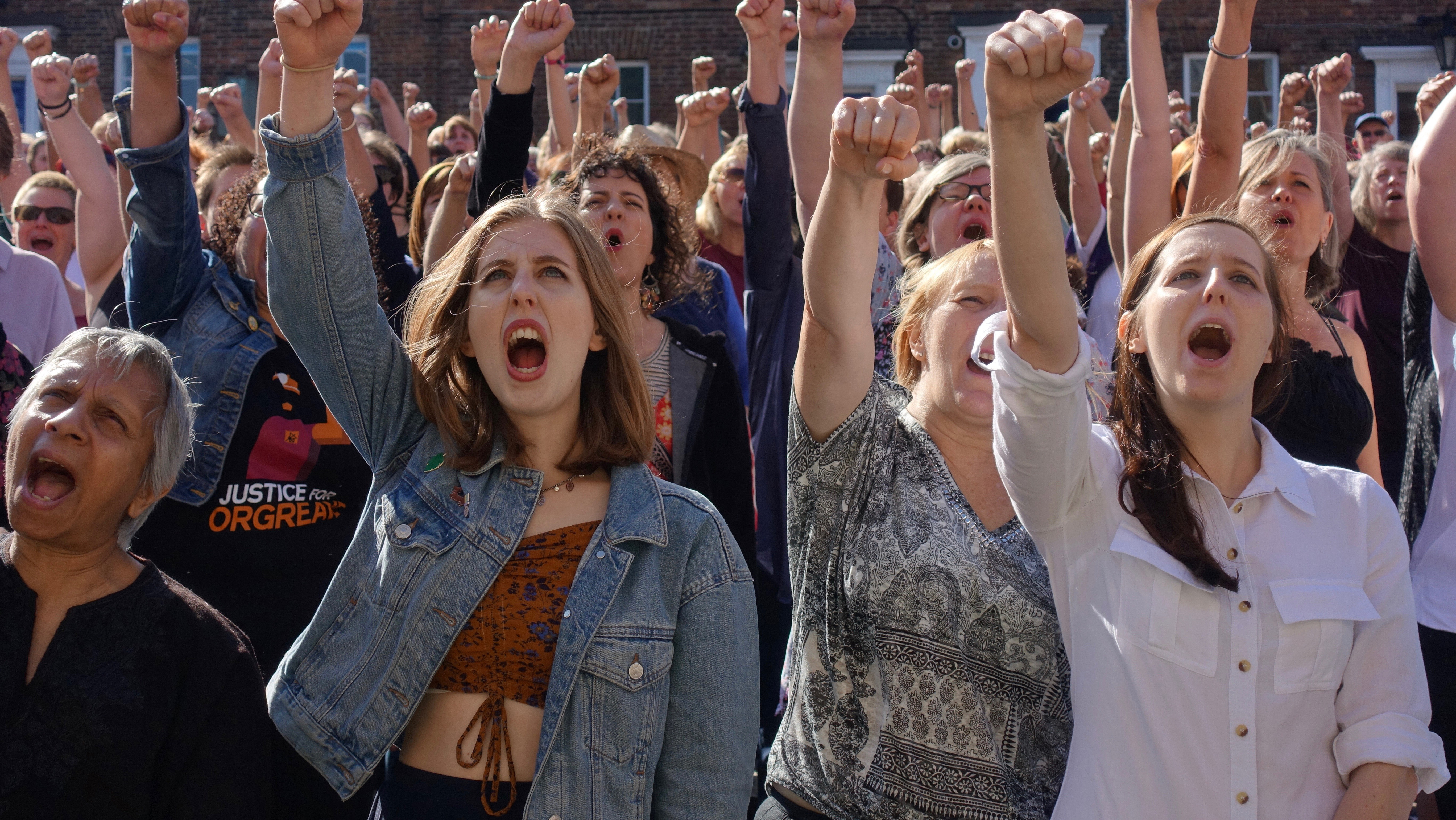 A photograph of a crowd of women at a protest or march. All have their mouths open, mid-shout. and have their right arms raised into a fist in the air. 
