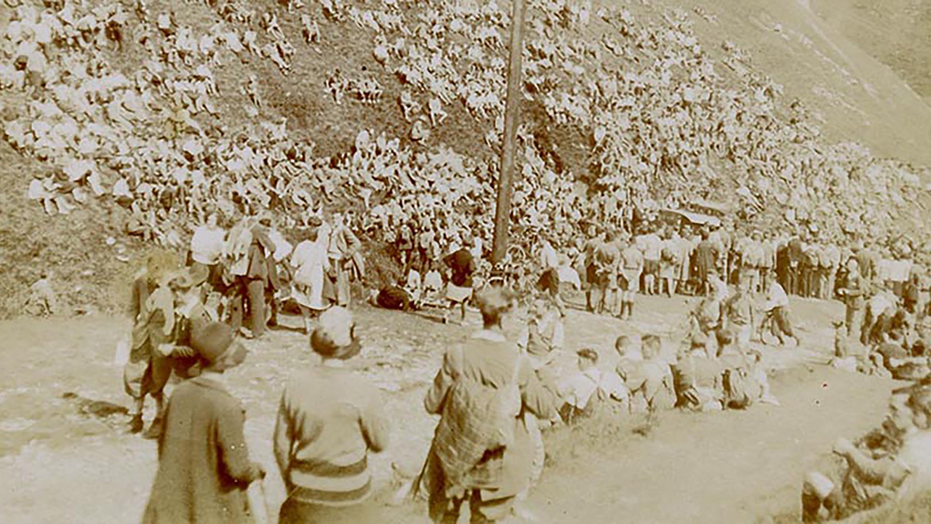 A sepia monochrome photo of a large crowd of adults and young people who are sat and stood on the hillside next to a large wooden pole.