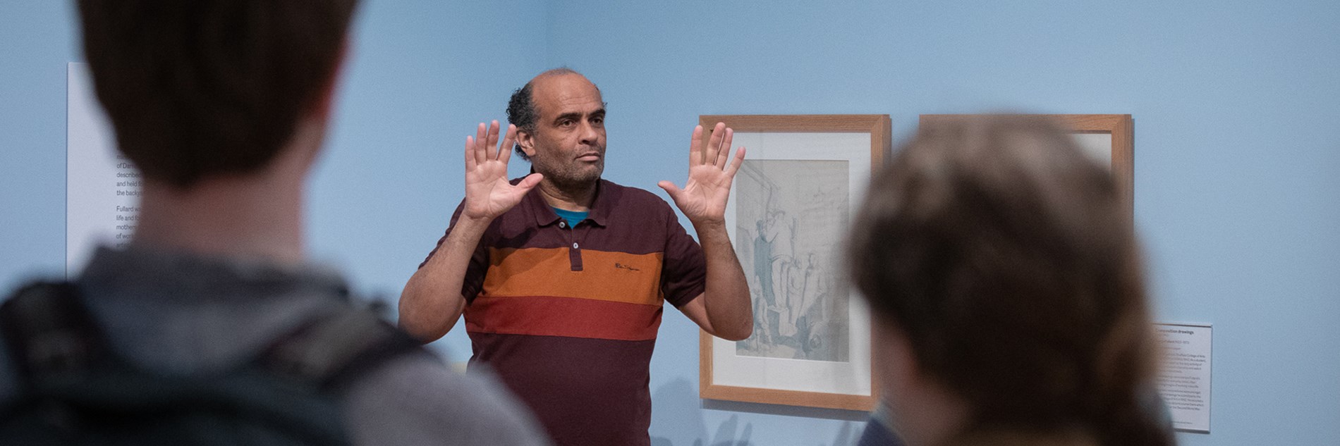 A photograph of an adult using British Sign Language. They are standing in front of a painting in an art gallery. 