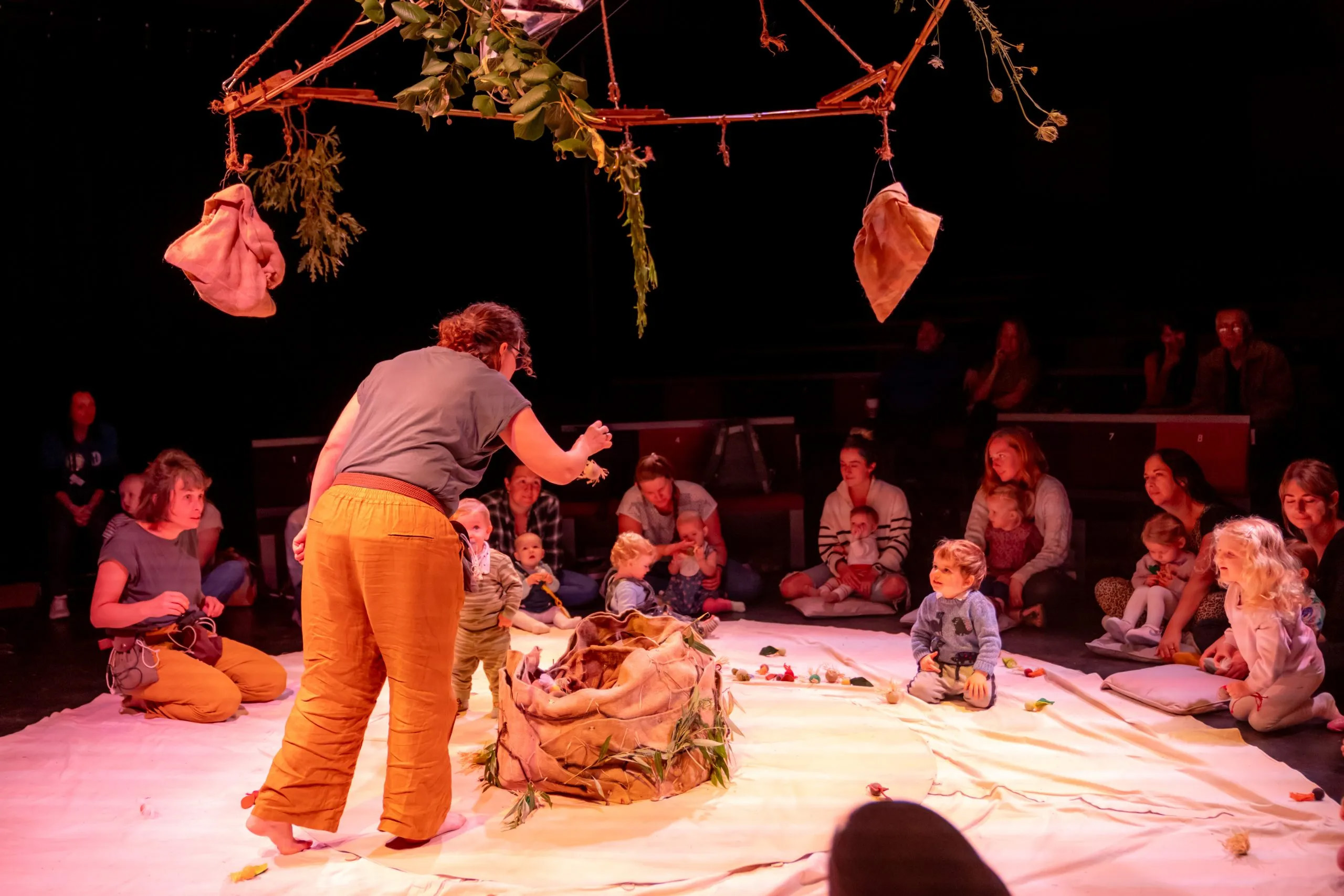 An actor on a stage in the round surrounded by an audience of infants with their parents.