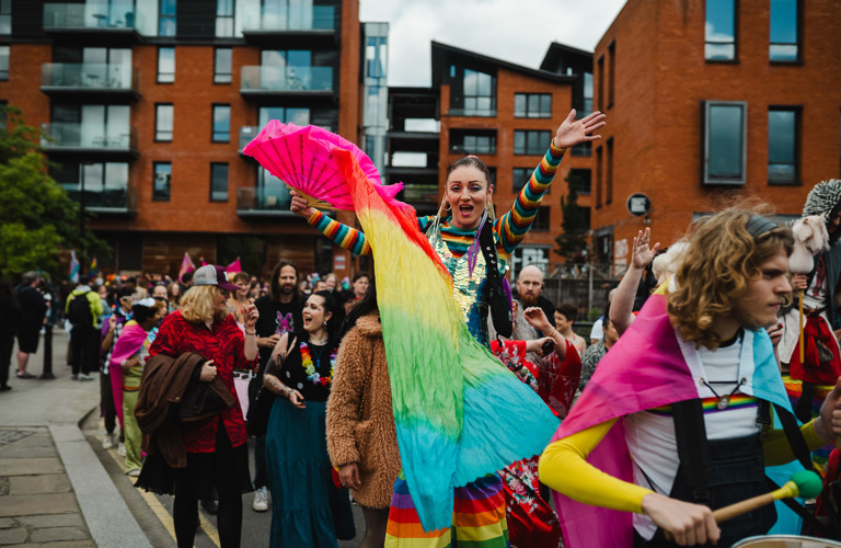 A photograph taken at a pride parade. The focal point of the picture is a young female-presenting person dressed in rainbow colours, holding a rainbow flag and lifting both arms in the air in celebration. A parade of many people continues around her.