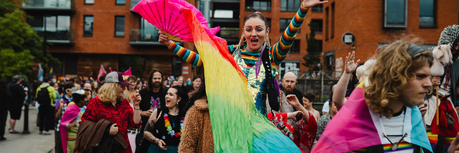 A photograph taken at a pride parade. The focal point of the picture is a young female-presenting person dressed in rainbow colours, holding a rainbow flag and lifting both arms in the air in celebration. A parade of many people continues around her.
