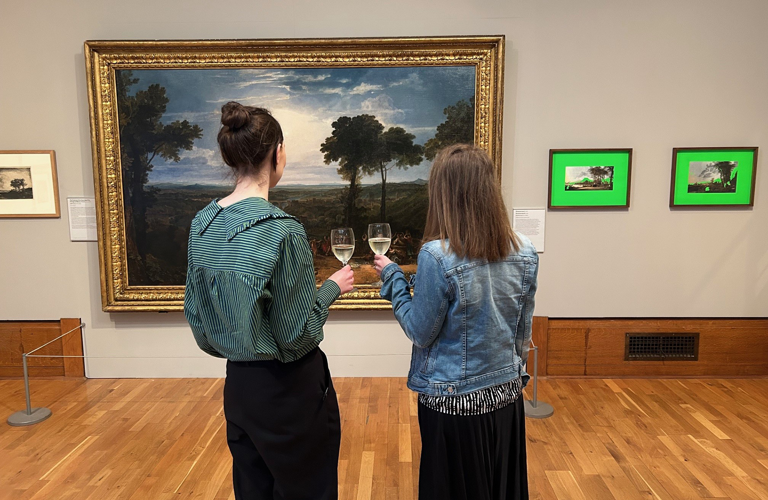 Two people looking at a painting in a gallery, holding wine glasses.