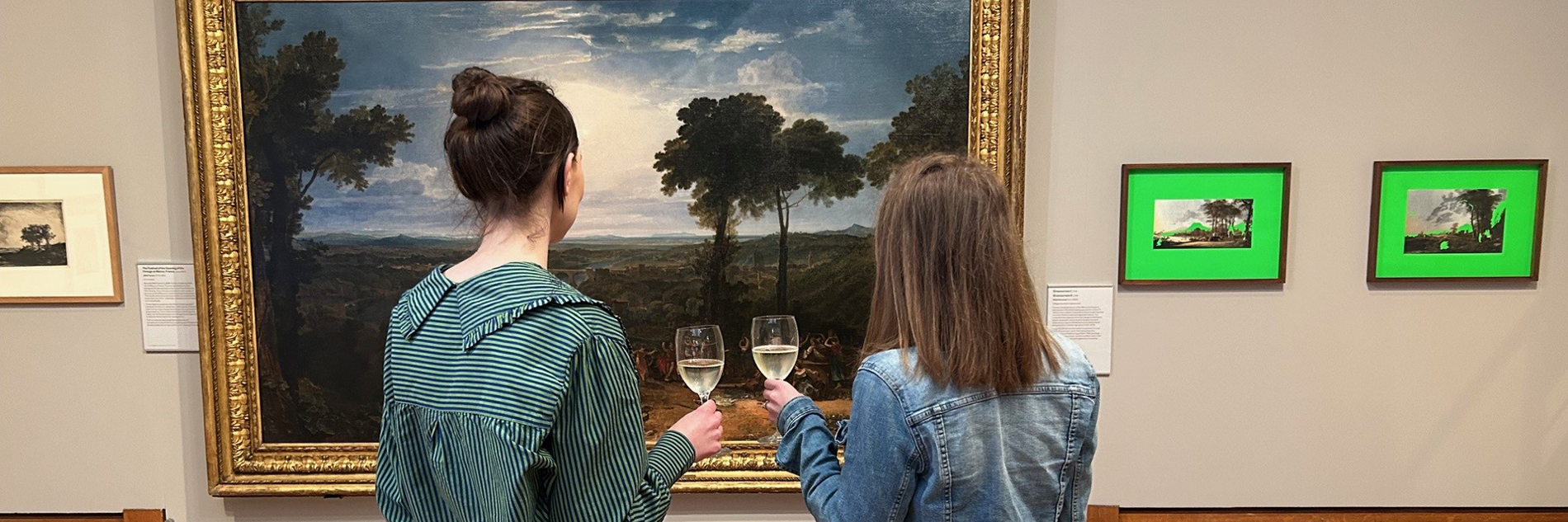 Two people looking at a painting in a gallery, holding wine glasses.