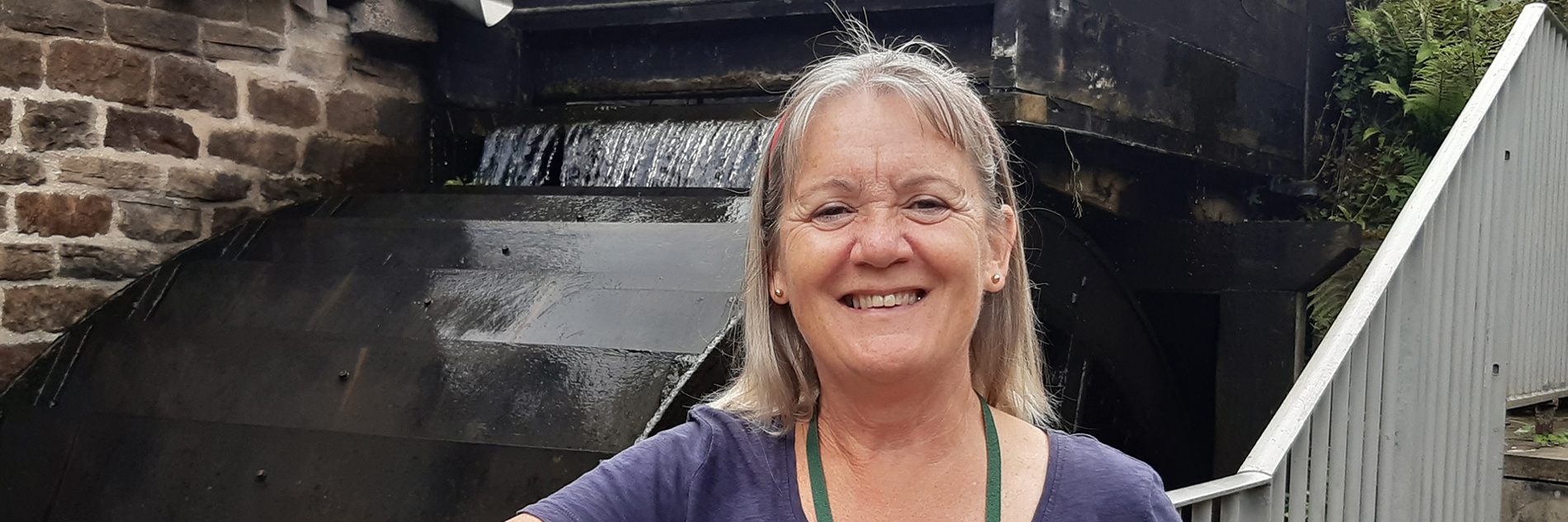 An adult stood in front of a metal handrail. Behind the handrail there is a historic running water wheel 