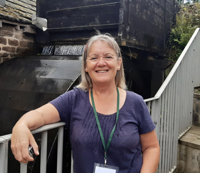 An adult stood in front of a metal handrail. Behind the handrail there is a historic running water wheel