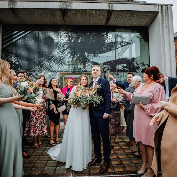 A bride in a wedding dress and a groom in a suit stand on front of a terrace in front of a contemporary building. Theye are surrounded by wedding guests.