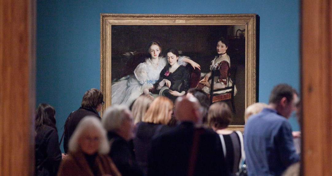 Adults in front of an oil painting, The Missus Vickers by John Singer Sargeant. The painting is of three women in formal Victorian dress. They are sitting informally next to each other - two women are sharing a chair and looking at a book, and the third woman is sitting on a chair behind them. The painting has a dark background, with the outline of a dresser on which sits a silver jug and a porcelain bowl. 
