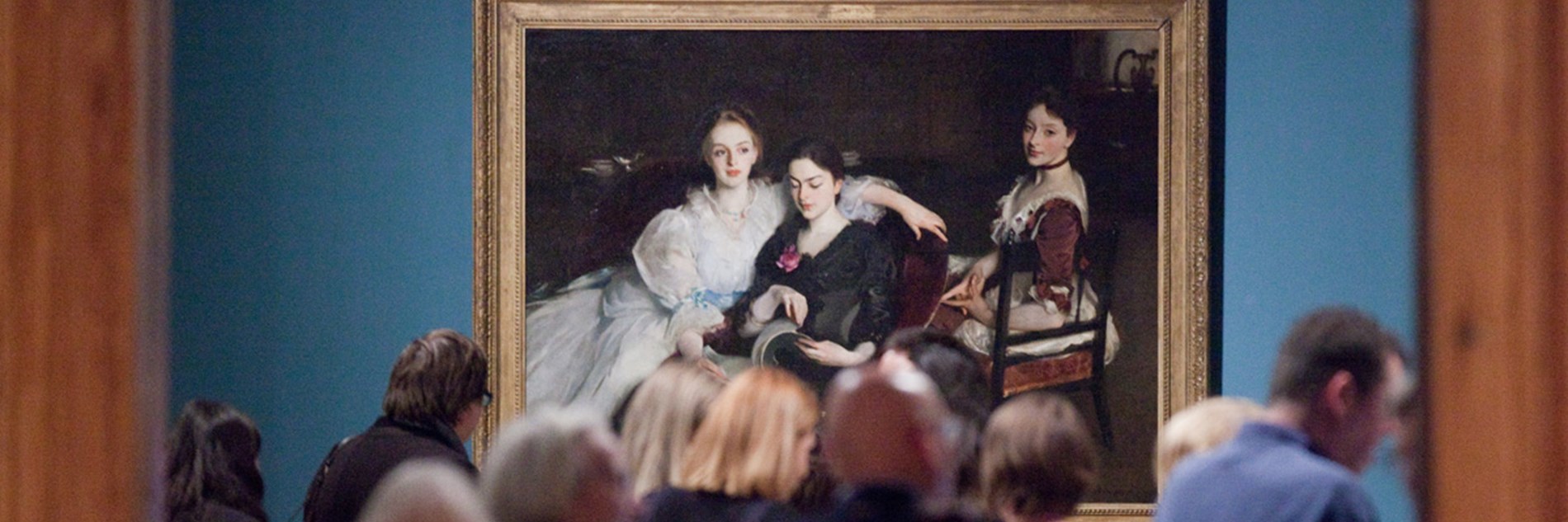 Adults in front of an oil painting, The Missus Vickers by John Singer Sargeant. The painting is of three women in formal Victorian dress. They are sitting informally next to each other - two women are sharing a chair and looking at a book, and the third woman is sitting on a chair behind them. The painting has a dark background, with the outline of a dresser on which sits a silver jug and a porcelain bowl. 