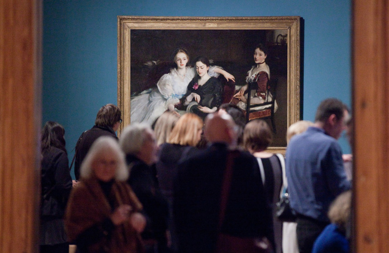 Adults in front of an oil painting, The Missus Vickers by John Singer Sargeant. The painting is of three women in formal Victorian dress. They are sitting informally next to each other - two women are sharing a chair and looking at a book, and the third woman is sitting on a chair behind them. The painting has a dark background, with the outline of a dresser on which sits a silver jug and a porcelain bowl. 