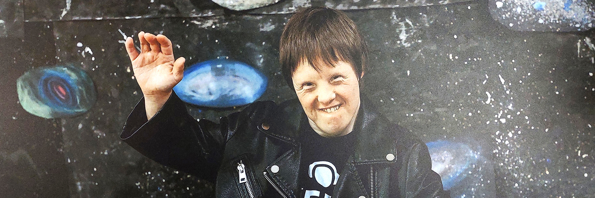 A smiling person stood behind DJ equipment waving. They stand in front of an abstract background that is reminiscent of space and stars.