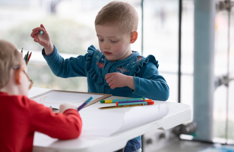 A photograph of a young child with cropped hair and a blue floral cardigan using felt tip pins to make a drawing