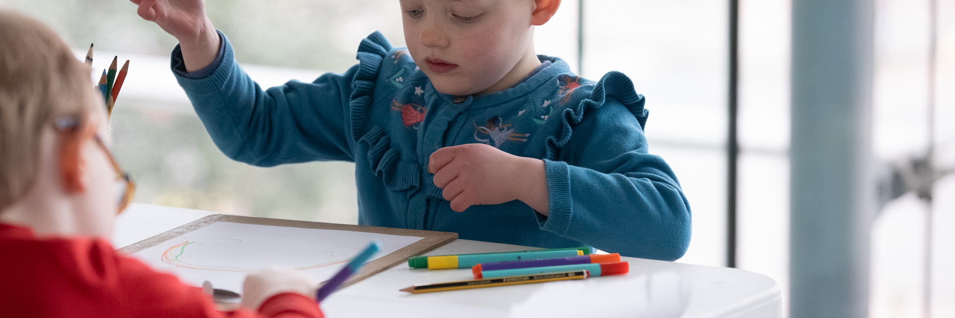 A photograph of a young child with cropped hair and a blue floral cardigan using felt tip pins to make a drawing