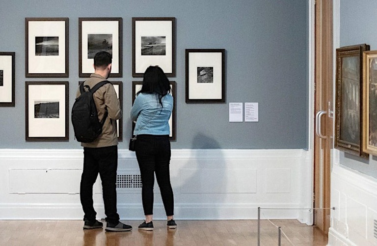 A photograph of two adults looking at a wall of black and white photographs in a gallery setting.